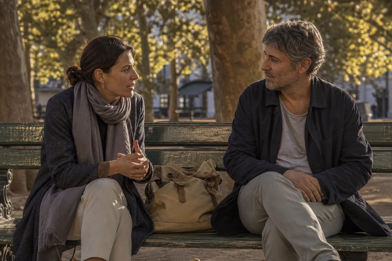 Two people sitting on a weathered park bench under plane trees, one speaking with hands still, the other listening, a canvas bag between them, dappled late-day light, 1600x900