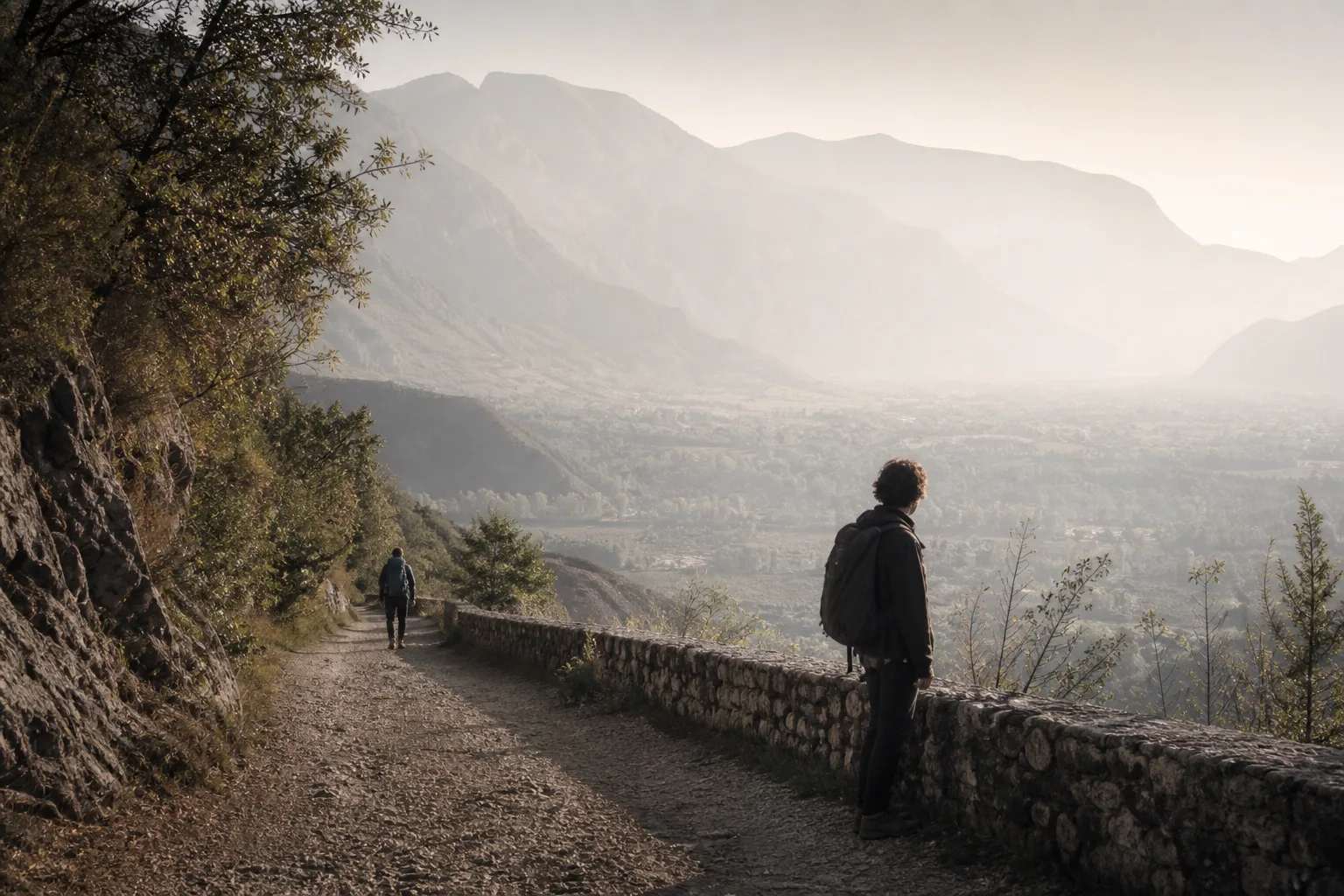 Two people walking the same gravel path at different distances, one far ahead with a daypack, one paused by a low stone wall looking at a valley, late morning haze, 1600x900